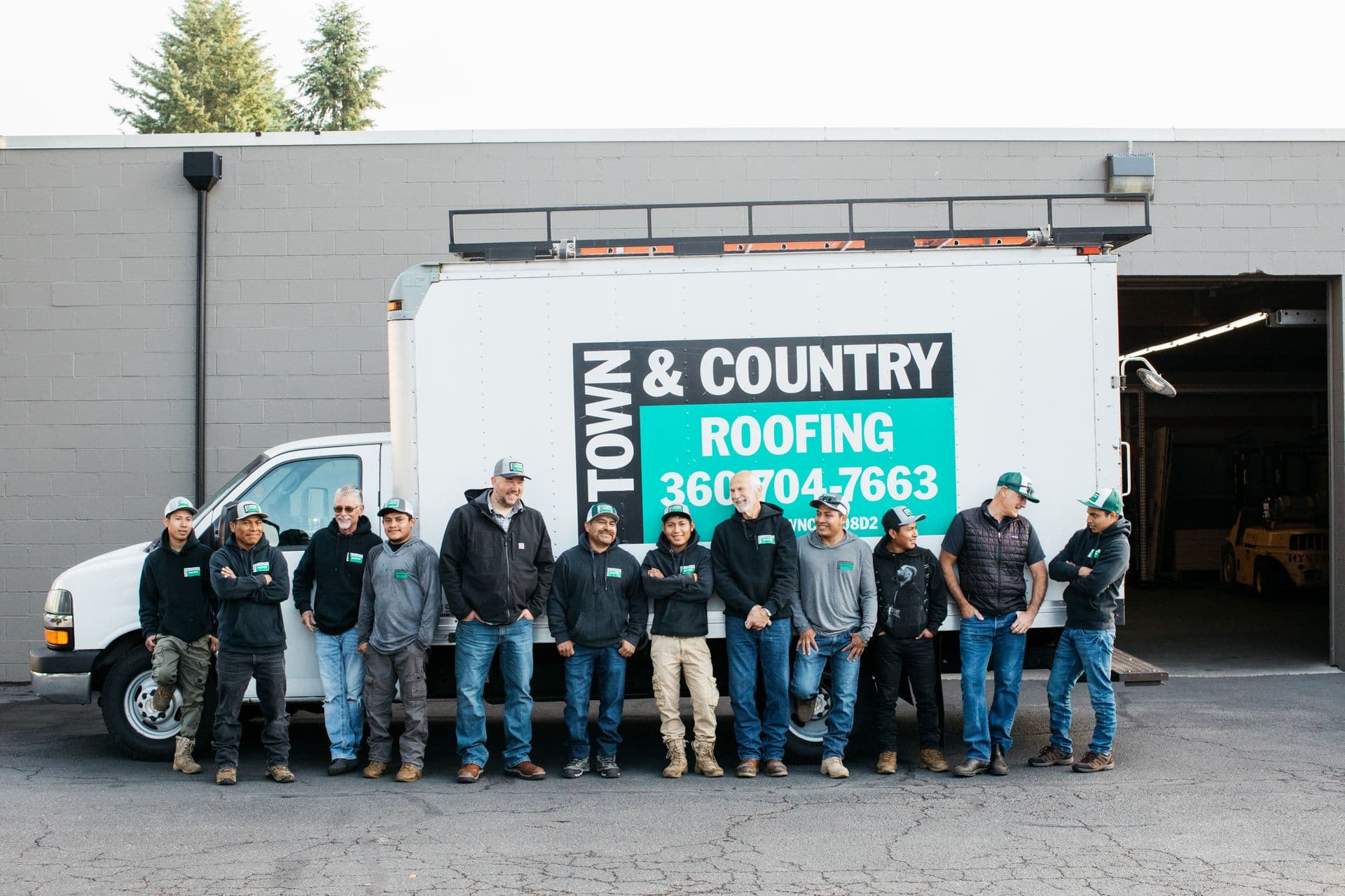 A team of eleven casually dressed workers stand smiling in front of a "Town & Country Roofing" truck. A "Master Shingle Applicator" sign is visible.