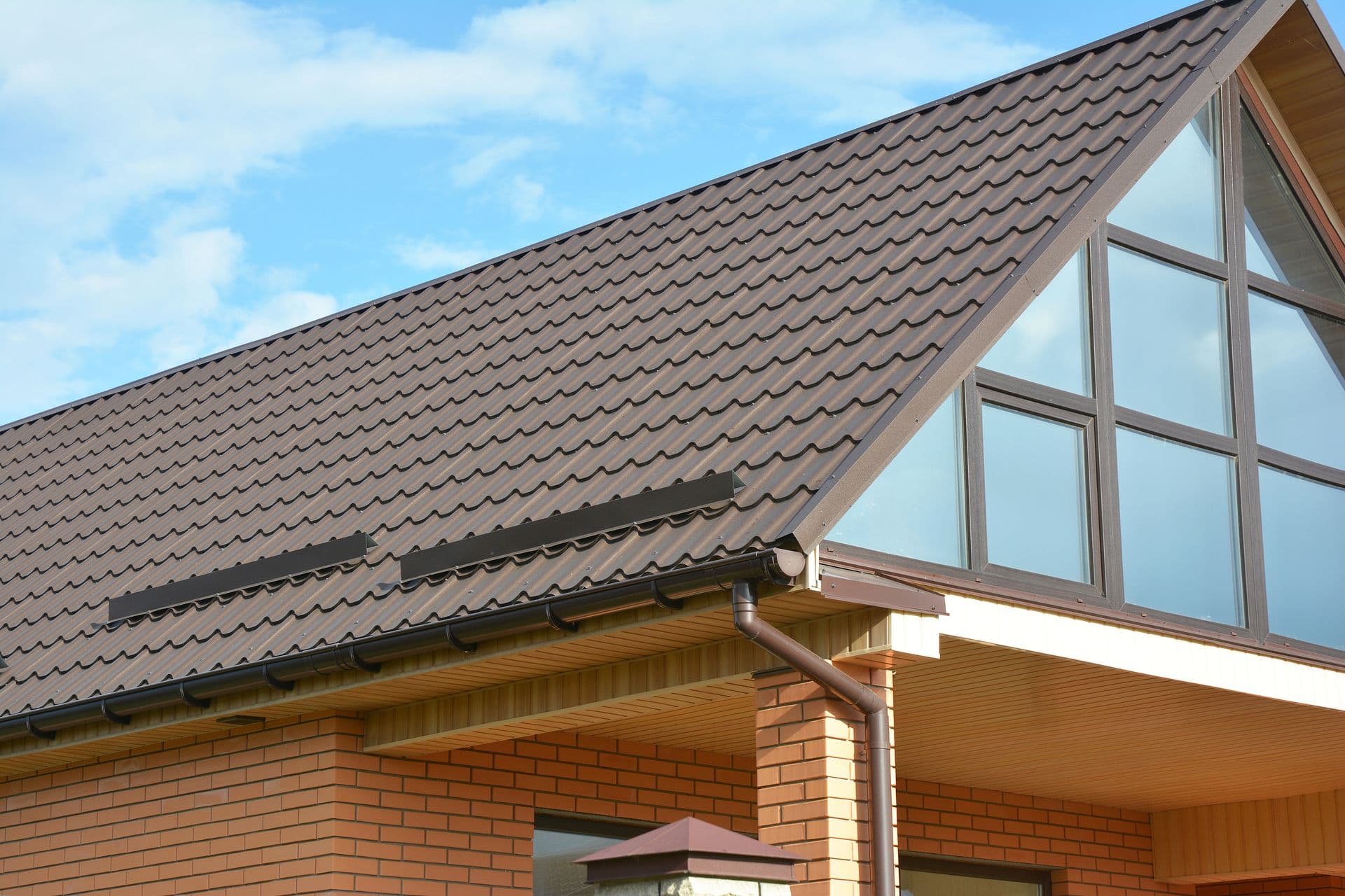 Gable roof with brown metal tiles on a brick house, featuring large glass windows and blue sky. The scene is bright and clear, conveying modern elegance.