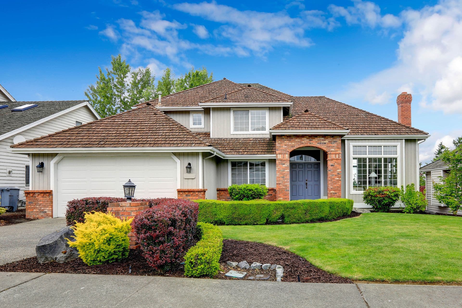 Two-story suburban house with a brown roof and brick accents. Manicured lawn with colorful shrubs. Bright blue sky and fluffy clouds overhead.