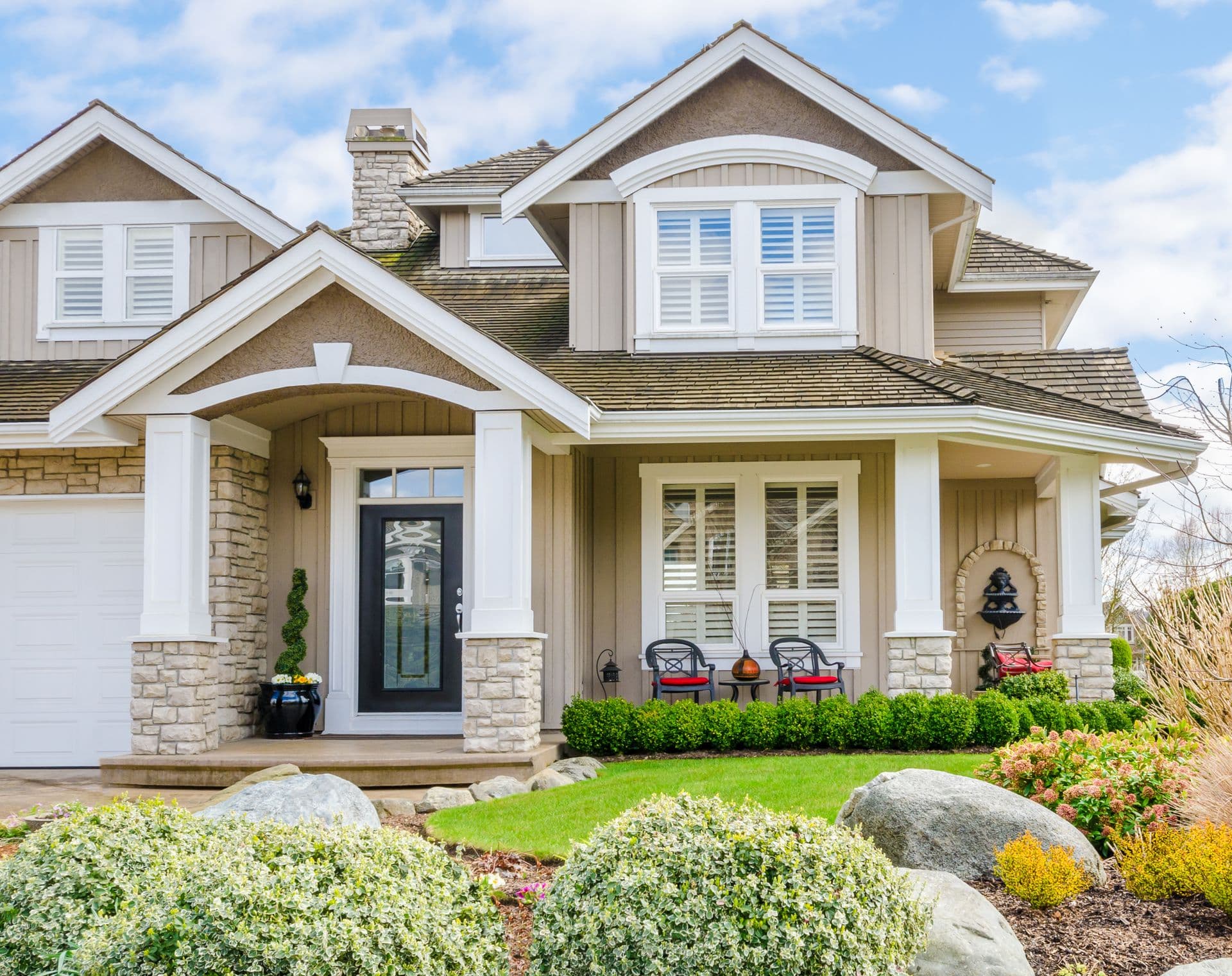 Charming suburban house with beige siding, white trim, and stone accents. Front porch has two black chairs with red cushions, surrounded by lush greenery.