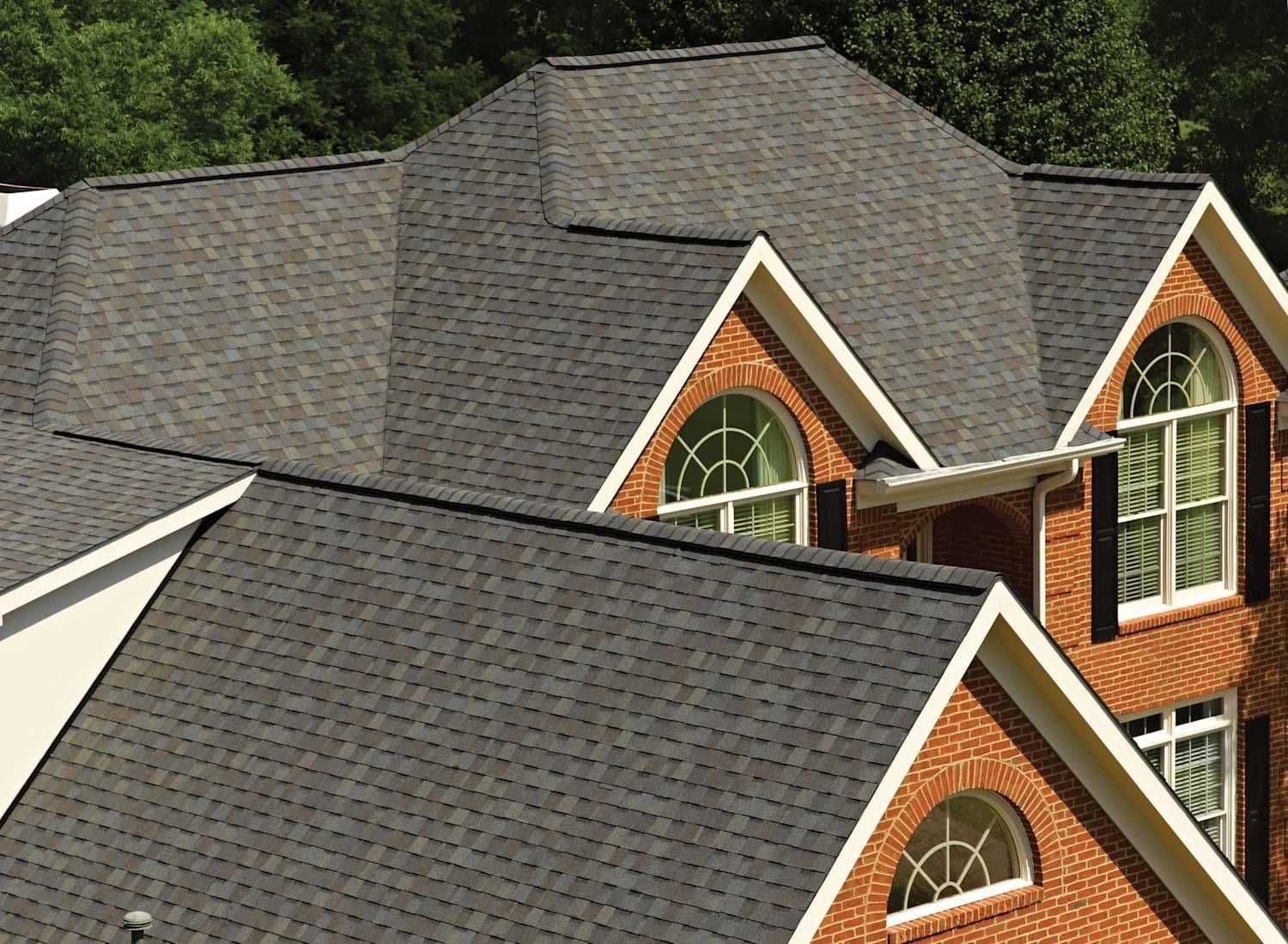 Alt text: A large, brick house with multiple steep gable roofs covered in dark gray shingles. Arched windows and lush green trees in the background enhance the elegant appearance.