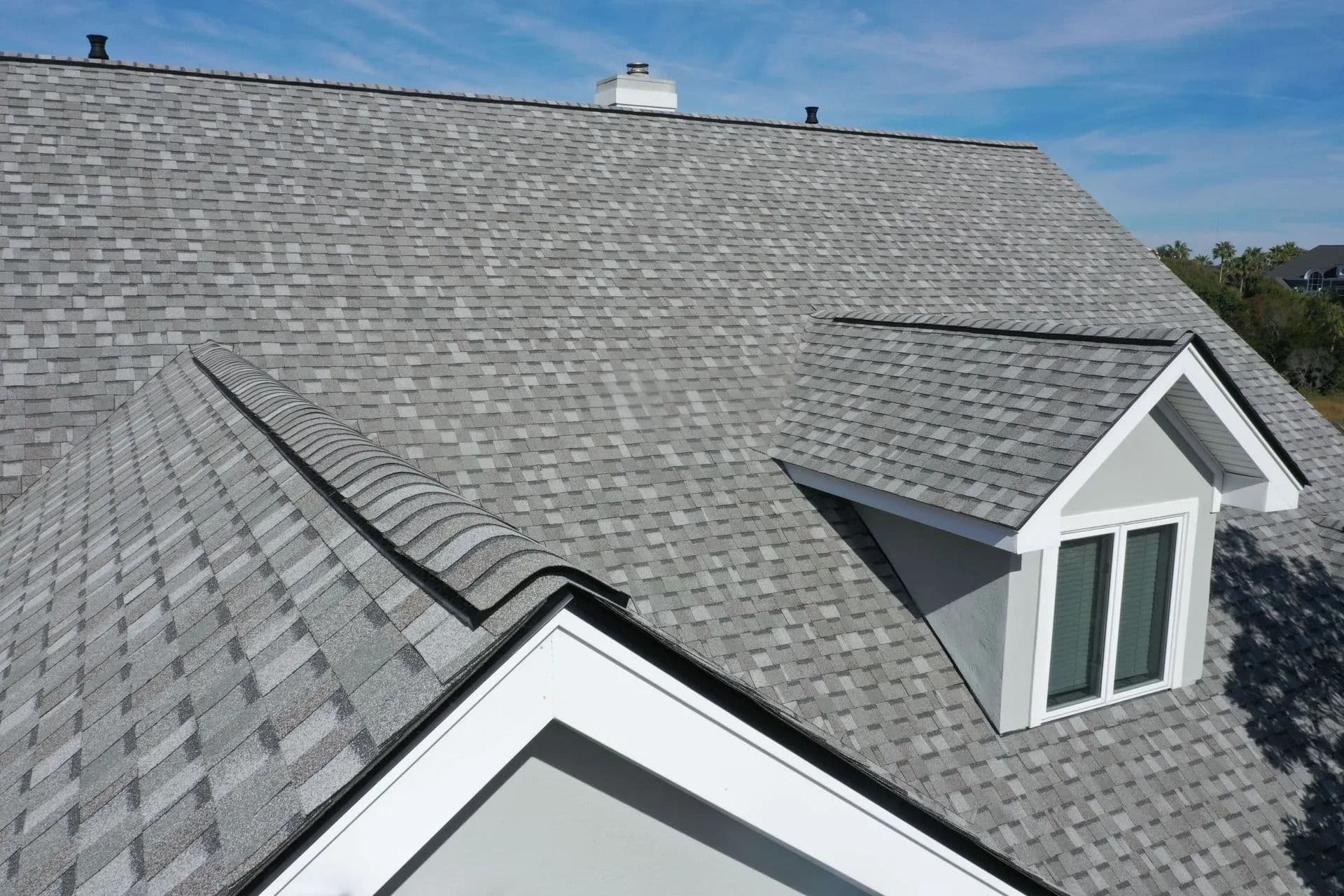 A roof with light gray shingles, featuring architectural details like dormer windows and white trim, under a bright blue sky with some clouds.