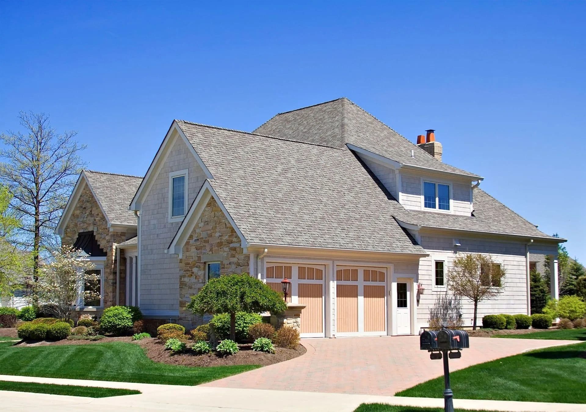A spacious modern house with beige siding and stone accents, featuring a three-car garage and manicured shrubs under a clear blue sky.
