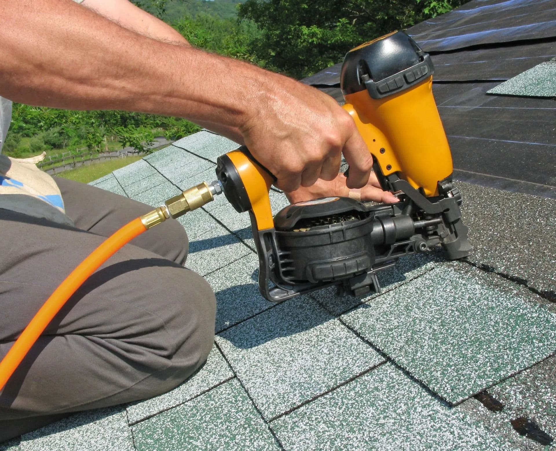 A person's hands using a yellow nail gun to install green asphalt shingles on a roof. The background shows a leafy landscape under clear skies.