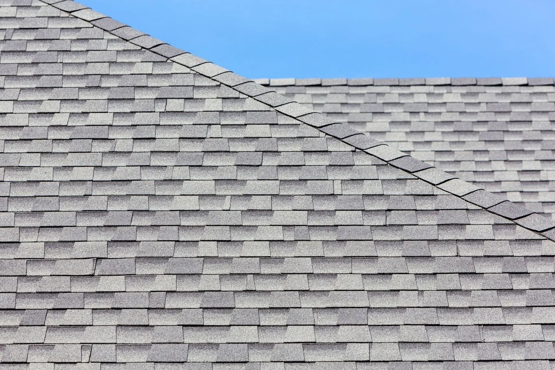 Close-up view of gray asphalt shingles on a residential roof against a clear blue sky. The pattern is uniform, conveying a sense of order and stability.