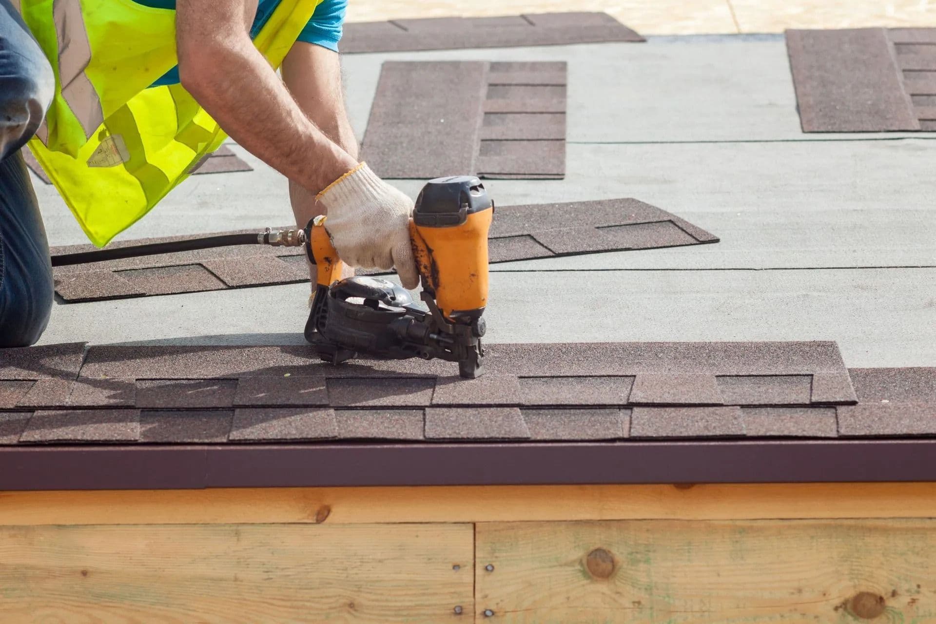 Worker in a high-visibility vest uses a nail gun to secure brown shingles on a roof. The focus is on precision and craftsmanship in roofing.