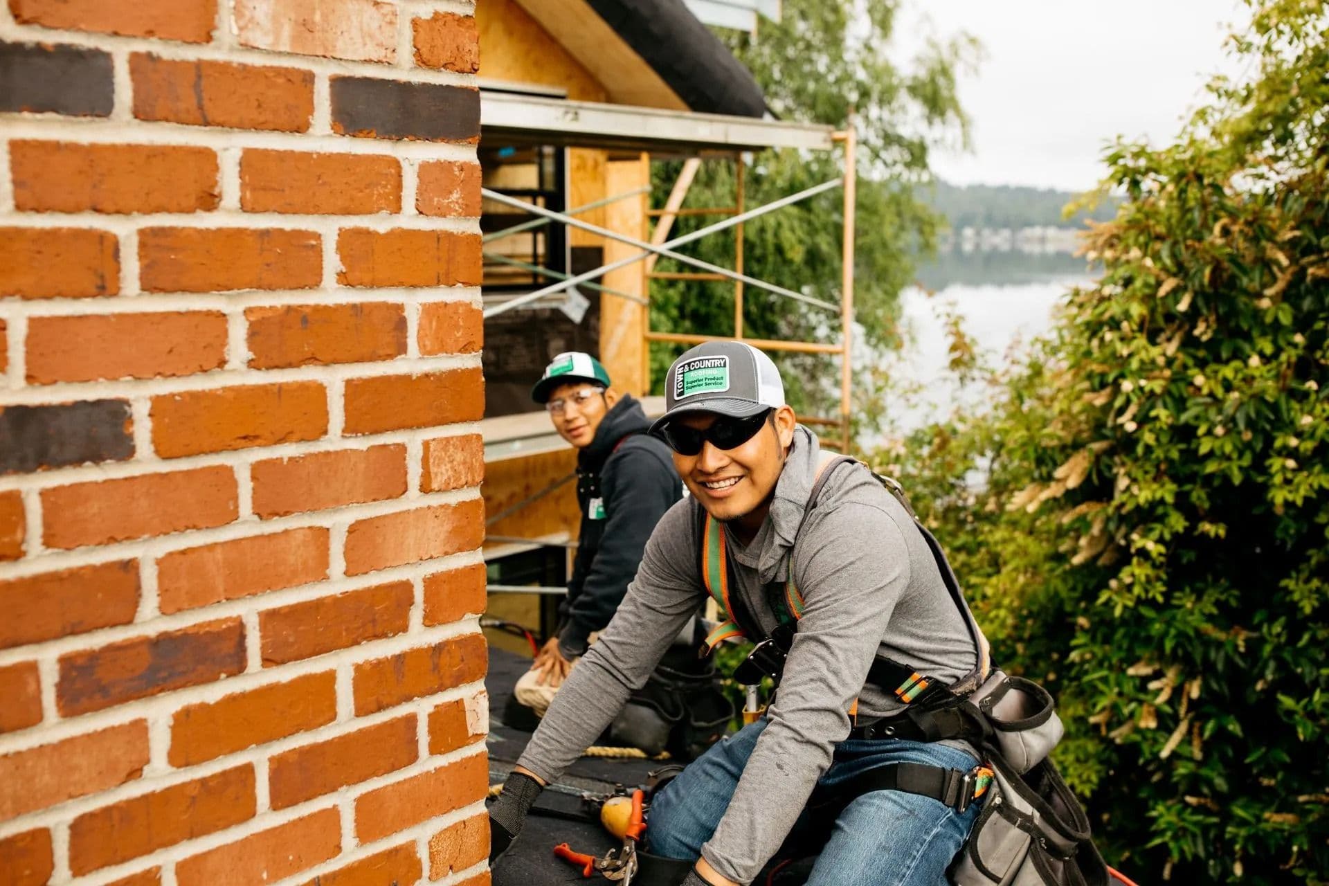 Two workers smiling on a rooftop near a brick chimney. They're wearing safety gear and caps, with greenery and a lake in the background.