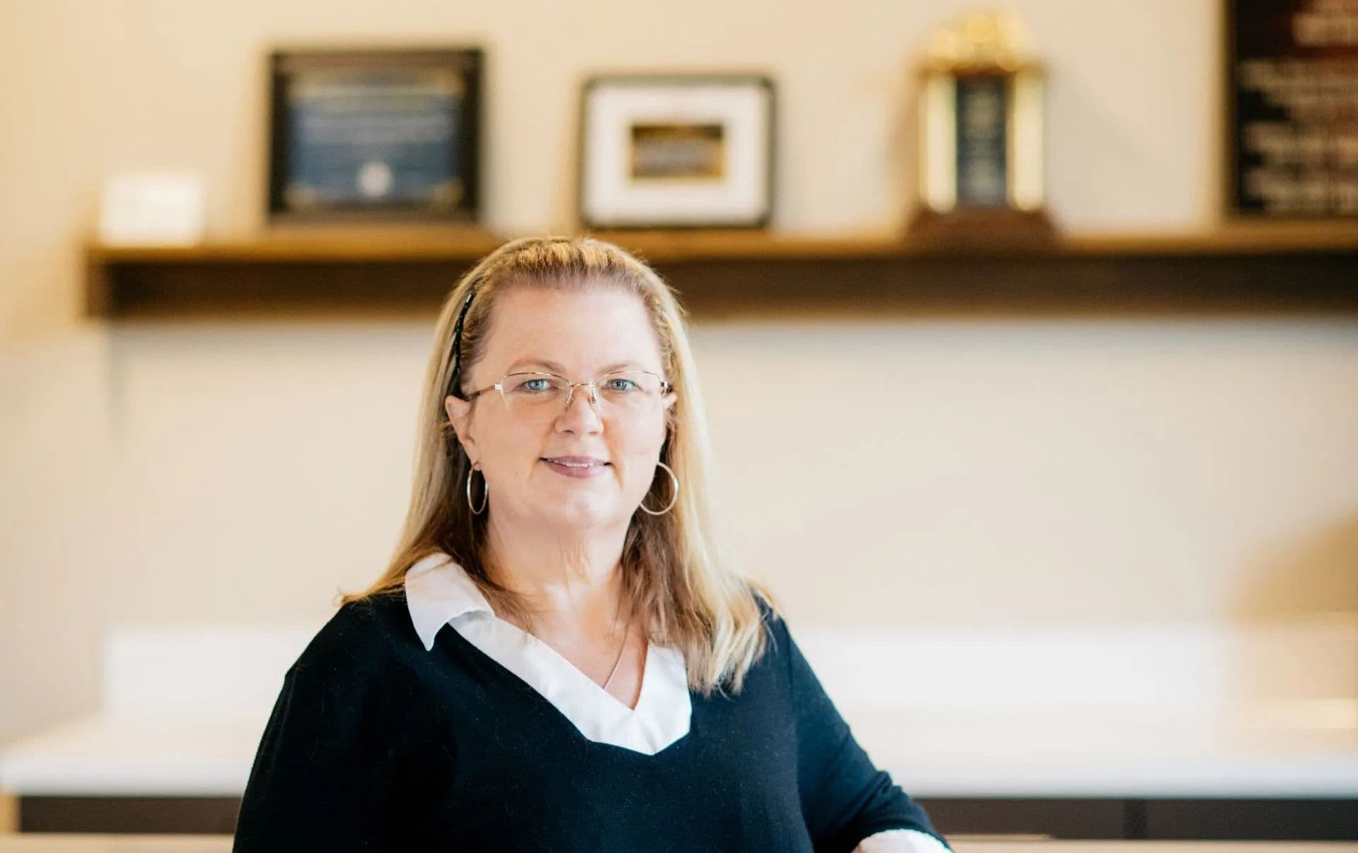 A woman with glasses and hoop earrings, wearing a black sweater over a white shirt, smiles in an office. Plaques and awards are blurred in the background.