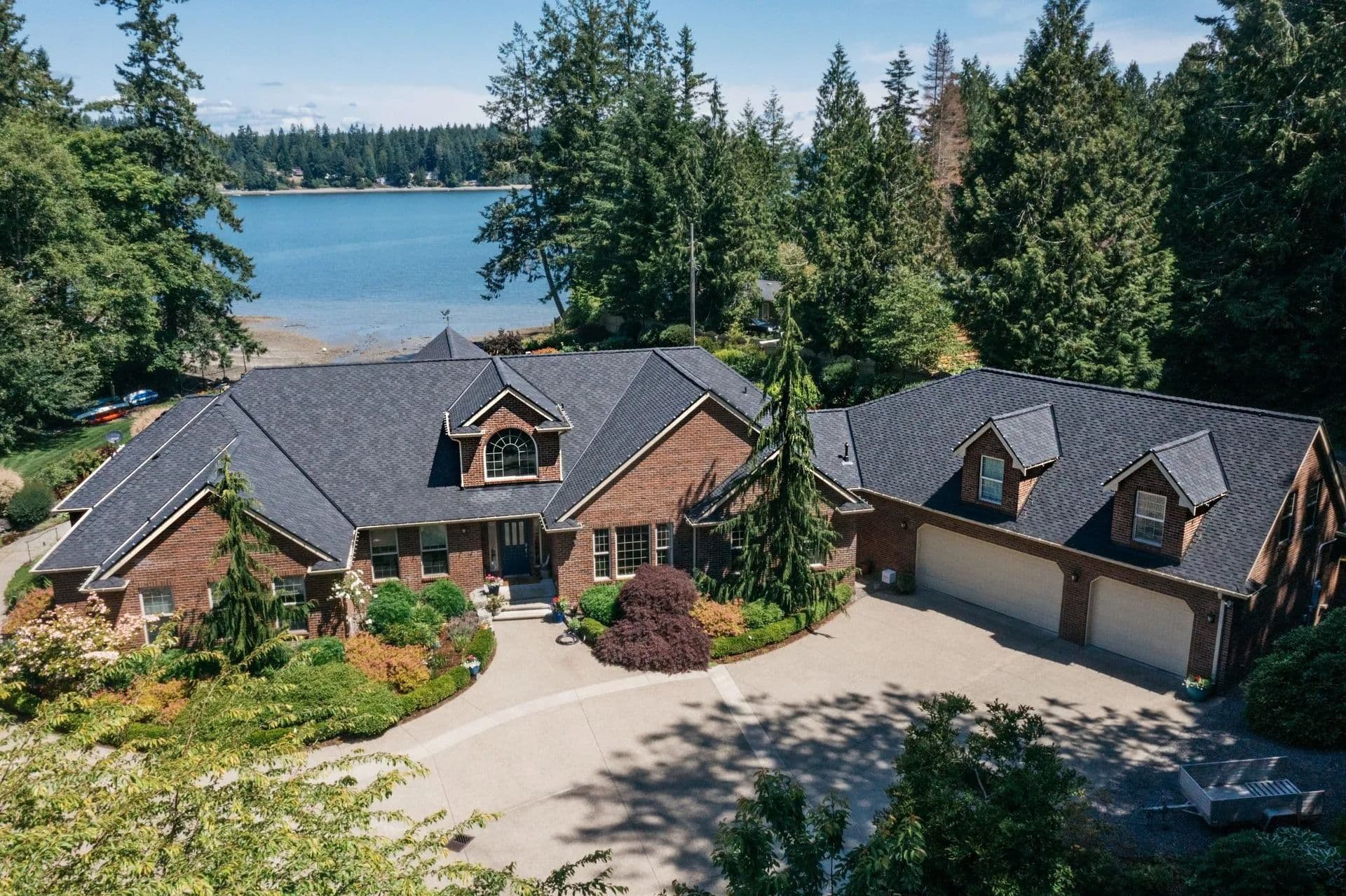 Aerial view of a large brick house with dark roofs, surrounded by lush trees and gardens. A calm blue lake is visible in the background under a clear sky.