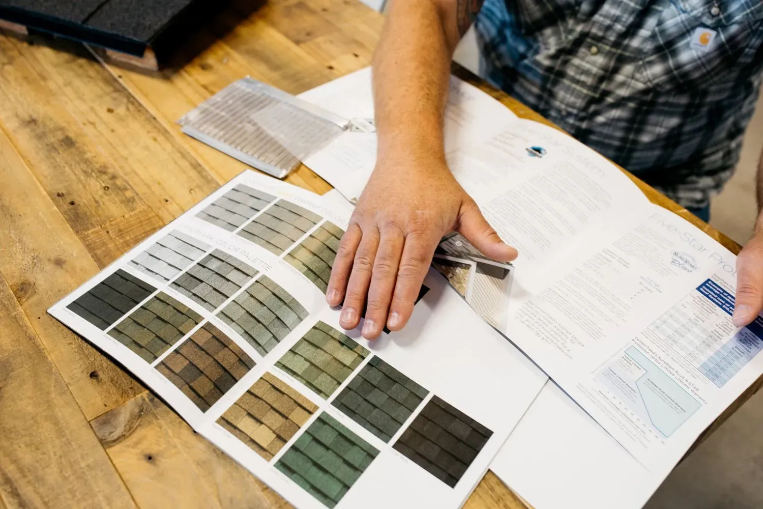 A person in a plaid shirt studies a roofing shingle color sample booklet on a wooden table, conveying a sense of thoughtful decision-making.