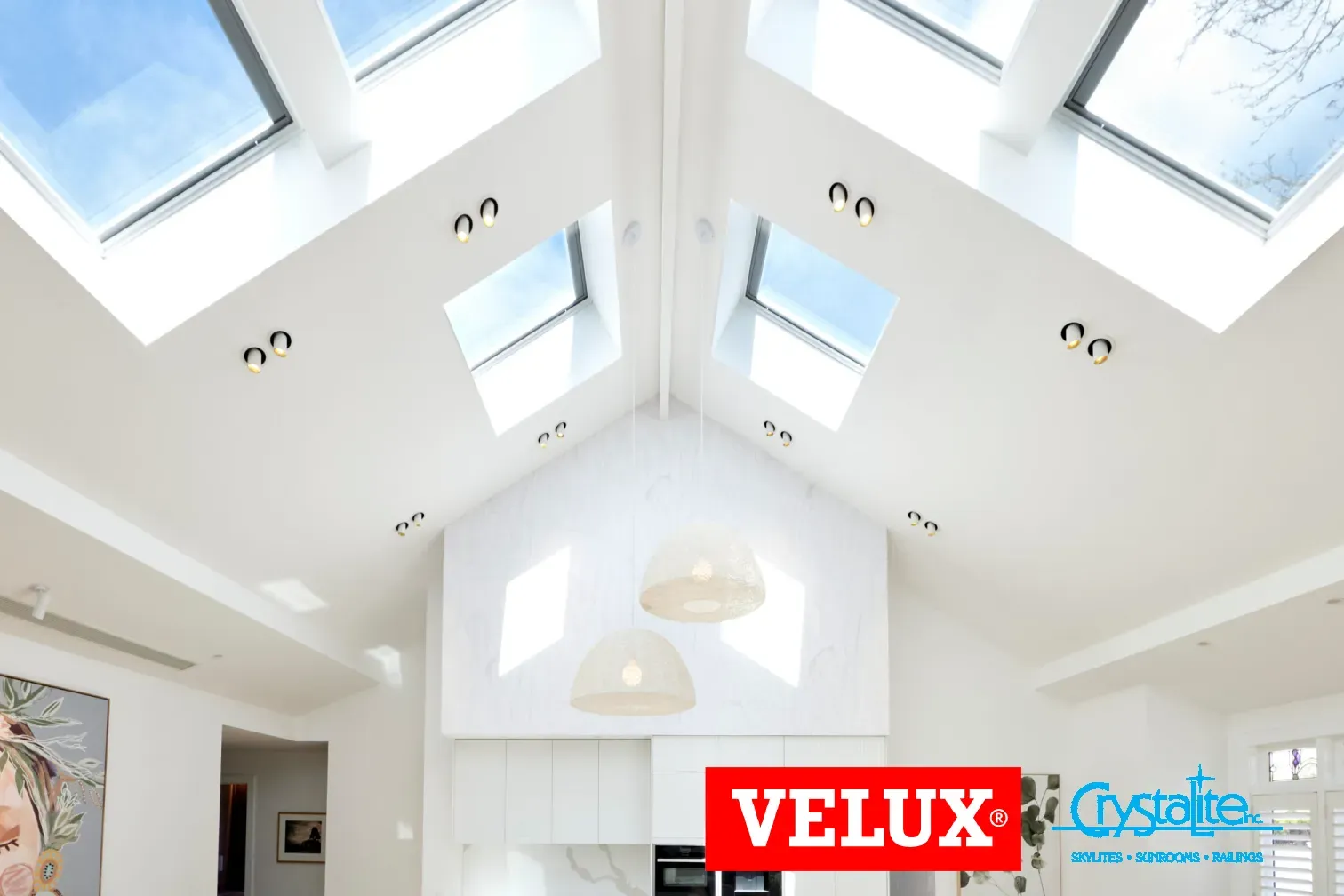 Bright, modern living space with large skylights on a high ceiling, white walls, hanging pendant lights, and a vibrant painting. Logos of VELUX and Crystalite are visible.