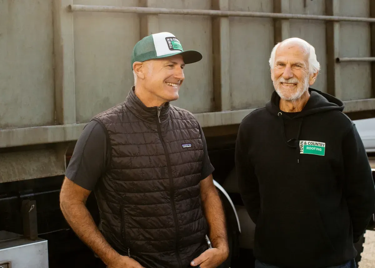 Two smiling men stand outdoors, one wearing a vest and cap, the other in a hoodie. They are beside a large industrial vehicle, conveying a friendly, relaxed mood.