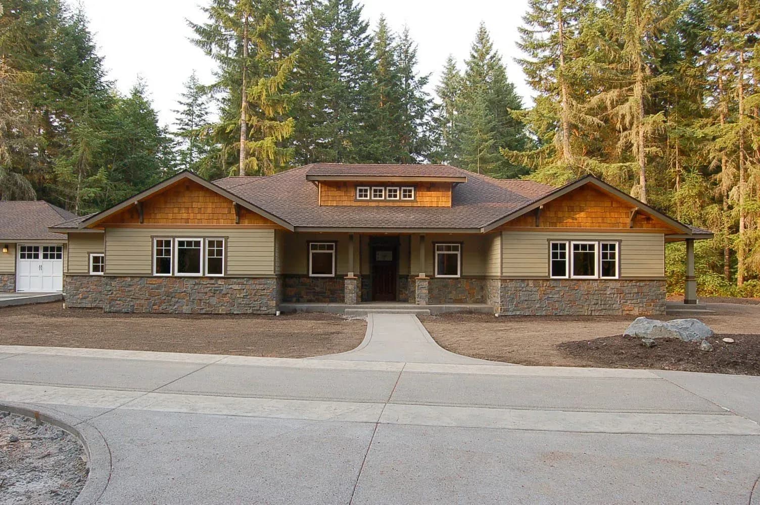 Single-story craftsman-style house with stone accents, surrounded by tall trees. A curved driveway leads up to the entrance, creating a serene setting.