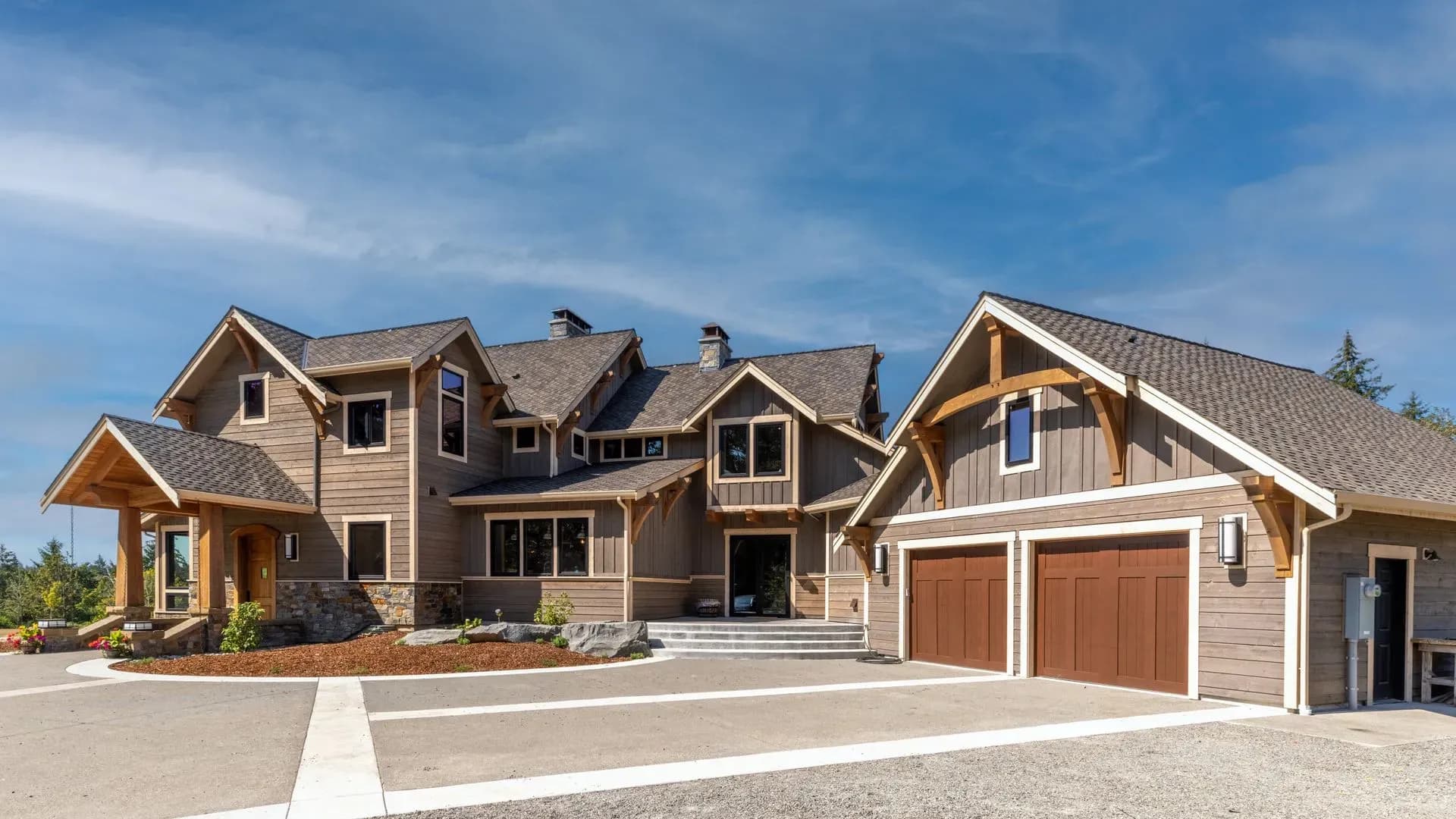 Spacious modern rustic house with wood and stone exterior, large windows, and a multi-car garage. Set against a clear blue sky, it conveys warmth and elegance.